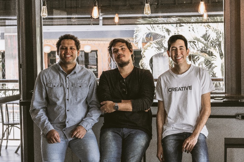 Three men are casually posing indoors. The man on the left is wearing a light denim shirt, the man in the middle is wearing a black long sleeve shirt and leaning against a counter, and the man on the right is wearing a white t-shirt with "CREATIVE" written on it.