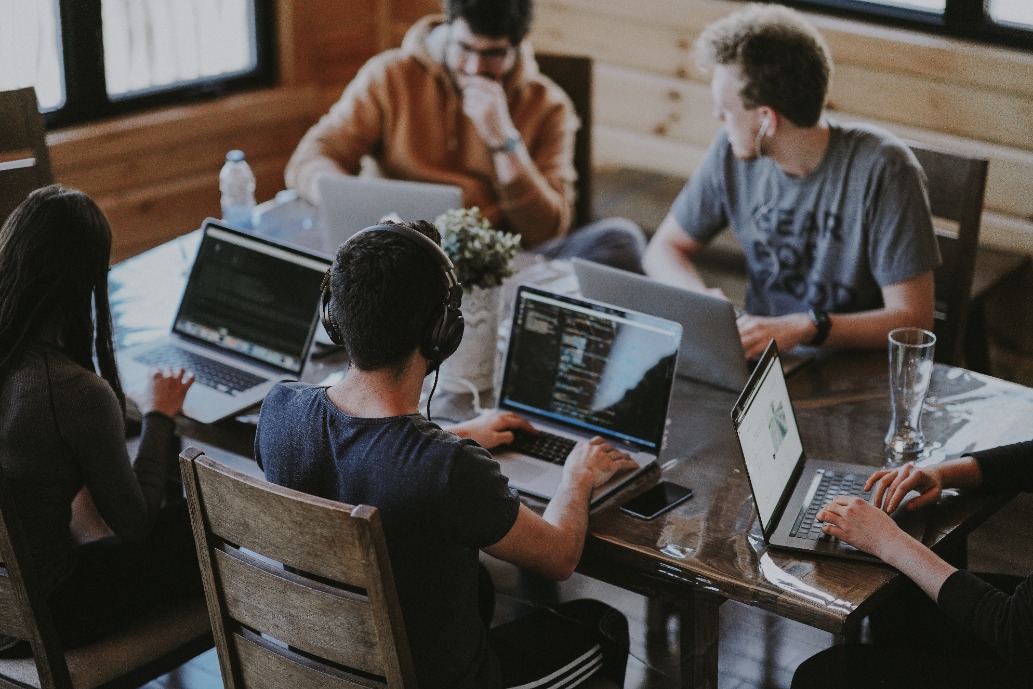 A group of people sit around a conference table in a modern, industrial-style room, working on laptops. A person stands at the front of the room, presenting a slide on a large screen. The room features wooden floors, concrete walls, and contemporary lighting.